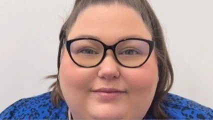 A person with long brown hair, wearing black-rimmed glasses and a blue top with black patterns, smiles softly at the camera against a plain light background.