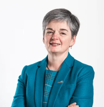 A woman with short grey hair wearing a teal blazer and checked top stands with arms crossed, smiling in front of a plain white background.