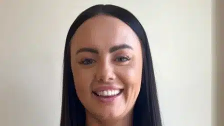 A woman with straight, dark brown hair and light skin is smiling at the camera. She is standing in front of a plain, light-coloured background.