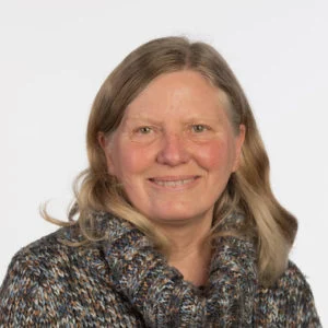 A middle-aged woman with light brown, shoulder-length hair smiles at the camera. She is wearing a thick, patterned jumper and is photographed against a plain white background.