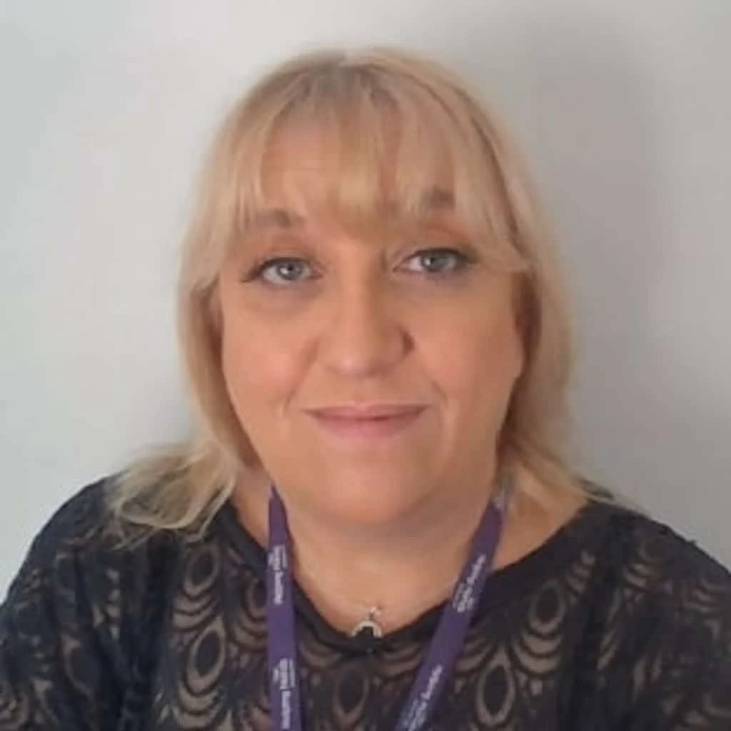 A woman with blonde hair and a fringe, wearing a black lace top and a purple lanyard, smiles softly at the camera against a plain light background.