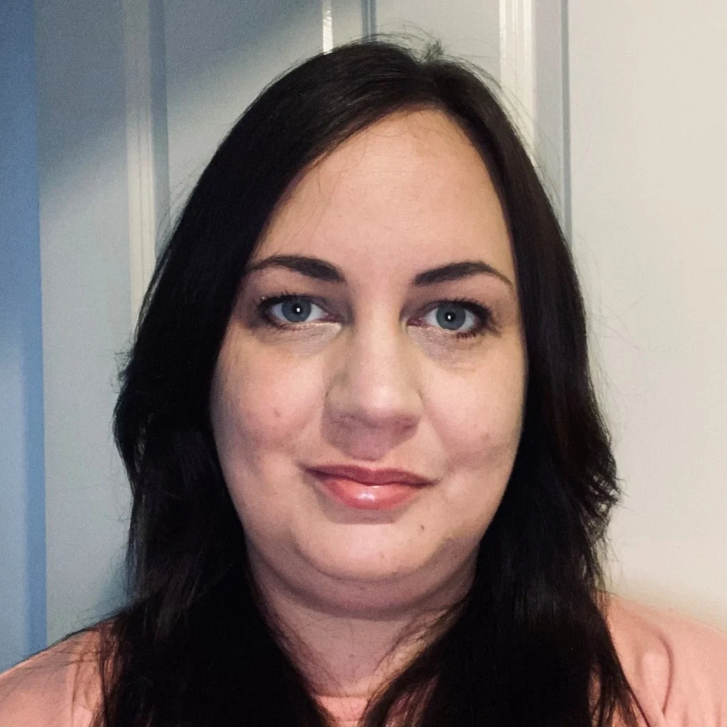 A woman with long dark hair and light eyes is standing in front of a white door, wearing a light pink top and looking directly at the camera with a slight smile.