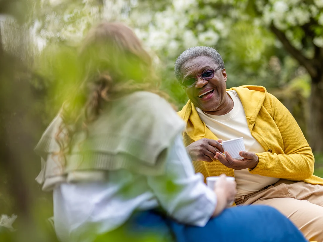 An older woman in a yellow jacket smiles and holds a cup while talking to another person outdoors, surrounded by greenery.