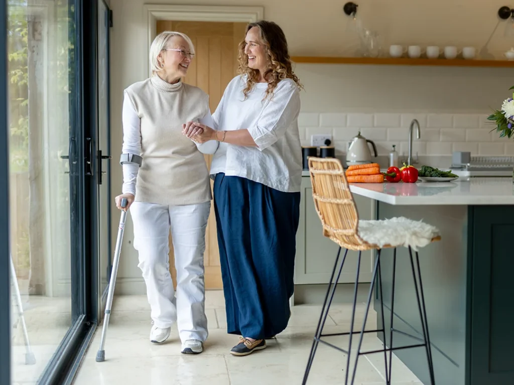Two women smile as they walk arm-in-arm in a bright kitchen. One uses a crutch for support. Vegetables and kitchen items are visible on the worktop, and a tall chair is in the foreground.