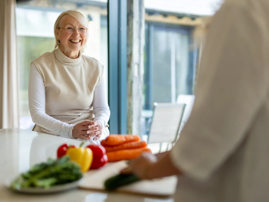 An older woman with glasses and fair hair sits at a kitchen island, smiling. Fresh vegetables, including peppers and carrots, are on the worktop. Another person is blurred in the foreground, preparing food.