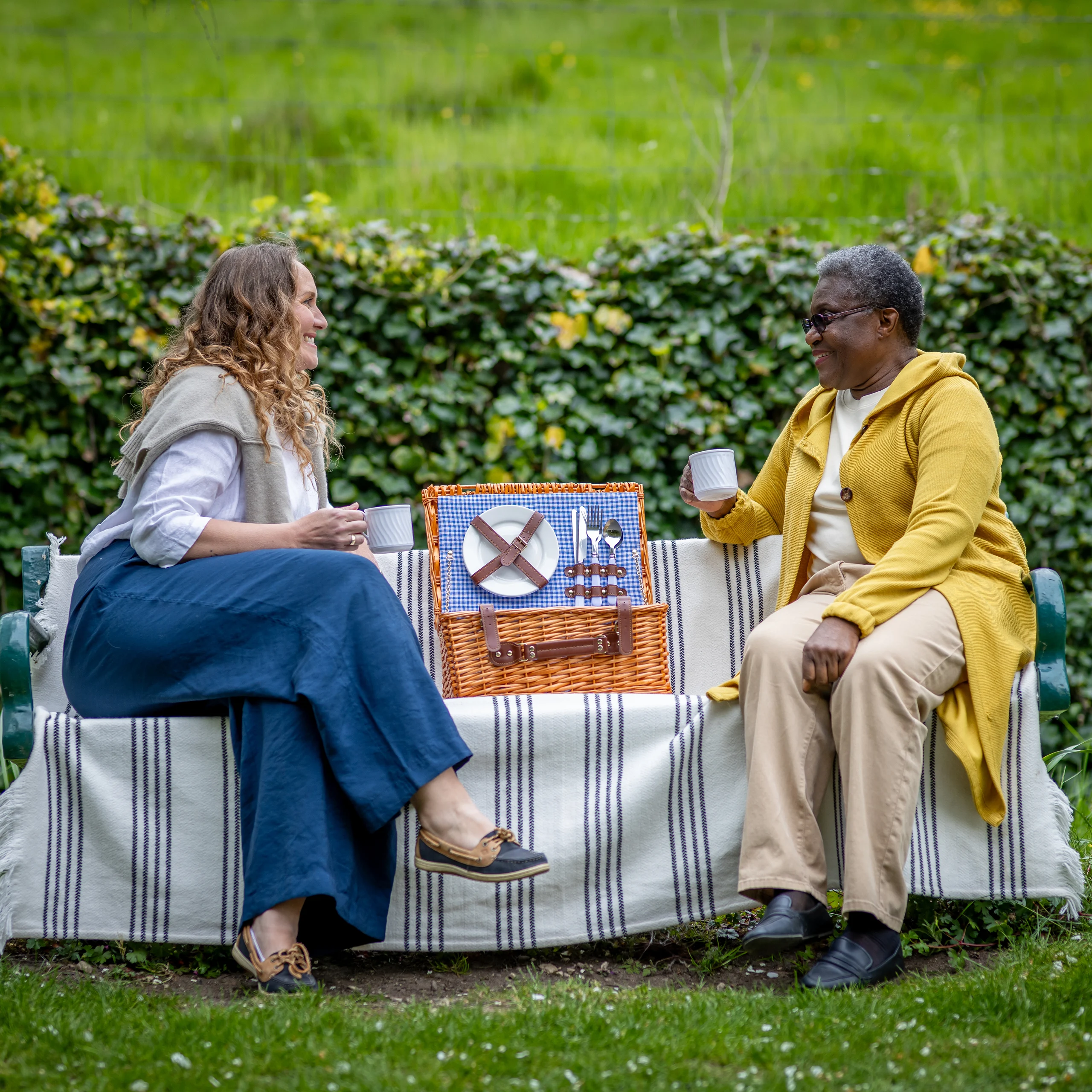 Two women sit on an outdoor bench covered with a striped blanket, enjoying drinks and smiling at each other. A picnic hamper with plates and cutlery is set between them. A hedge and green grass are in the background.