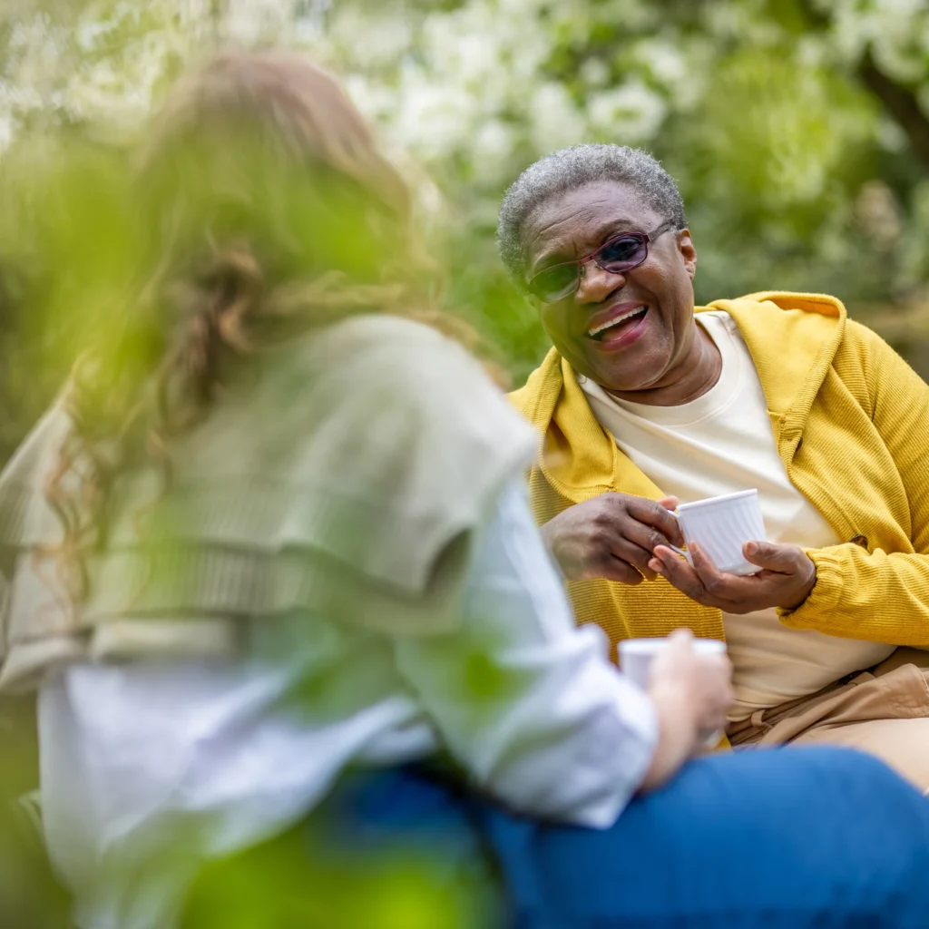 Two people sit outdoors, smiling and talking whilst holding cups. One person wears a yellow hoodie and glasses, and the other has long hair and a jumper over their shoulders. Green foliage surrounds them.