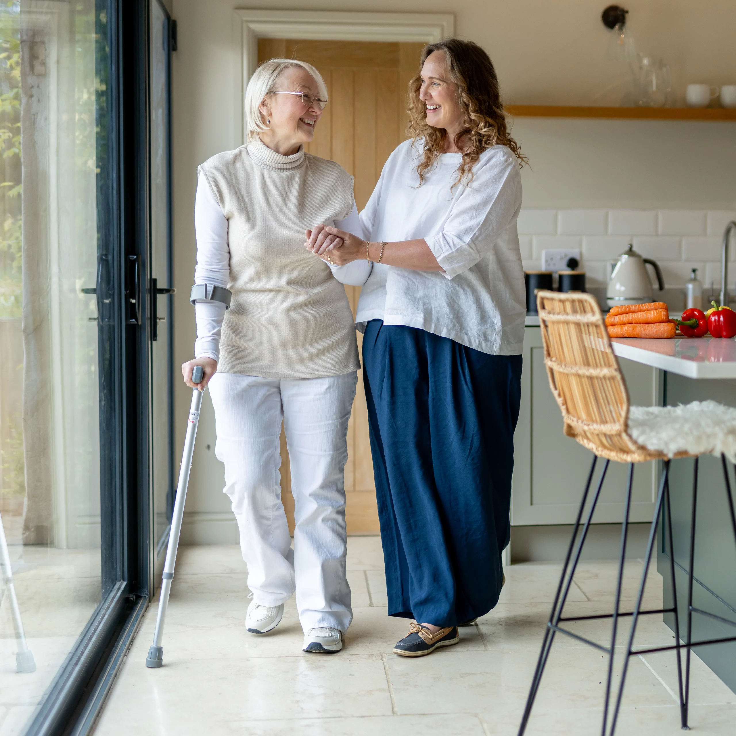 An older woman using a walking stick walks arm in arm with a younger woman in a bright kitchen, both smiling and looking at each other warmly.