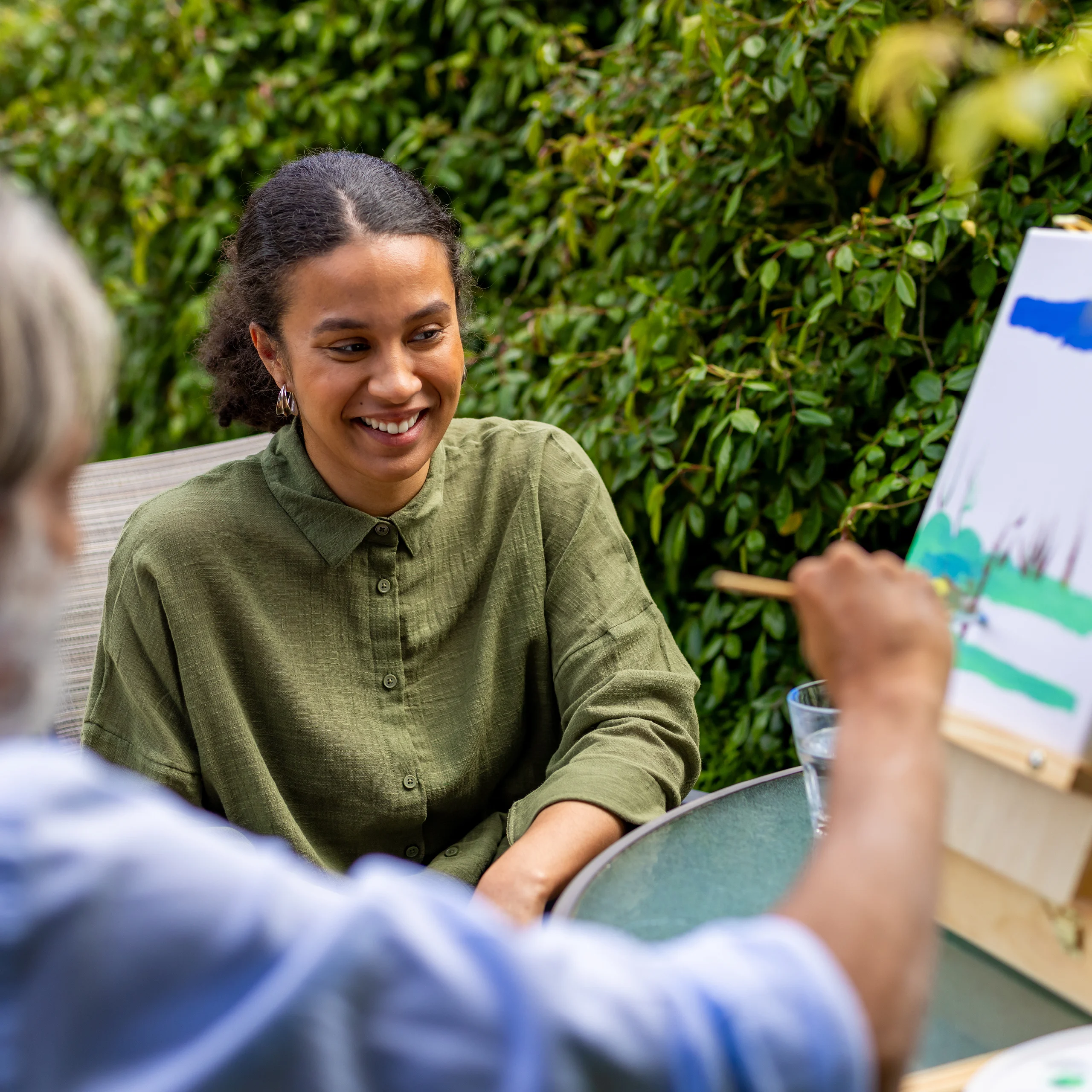 A young woman in a green shirt smiles whilst sitting at a glass table outdoors, watching another person paint on a small canvas with green and blue colours. Lush greenery fills the background.