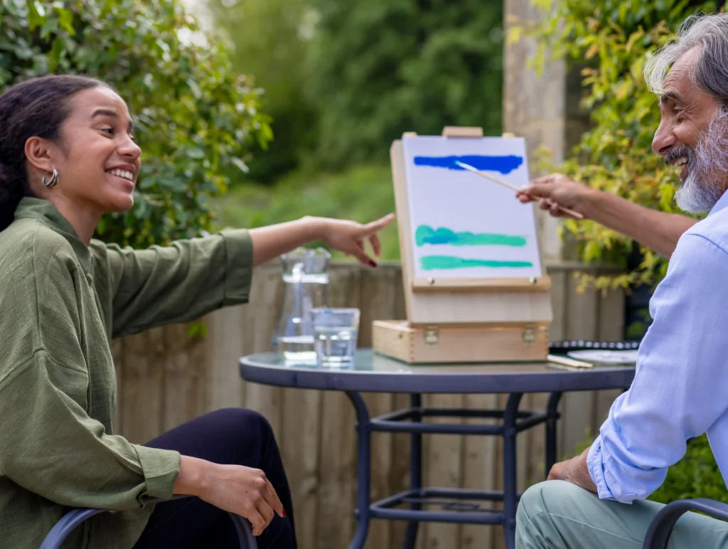 Two people sit outside at a round table, smiling and painting on a small easel. One person points at the canvas, while the other gestures with a paintbrush. There are glasses of water on the table.