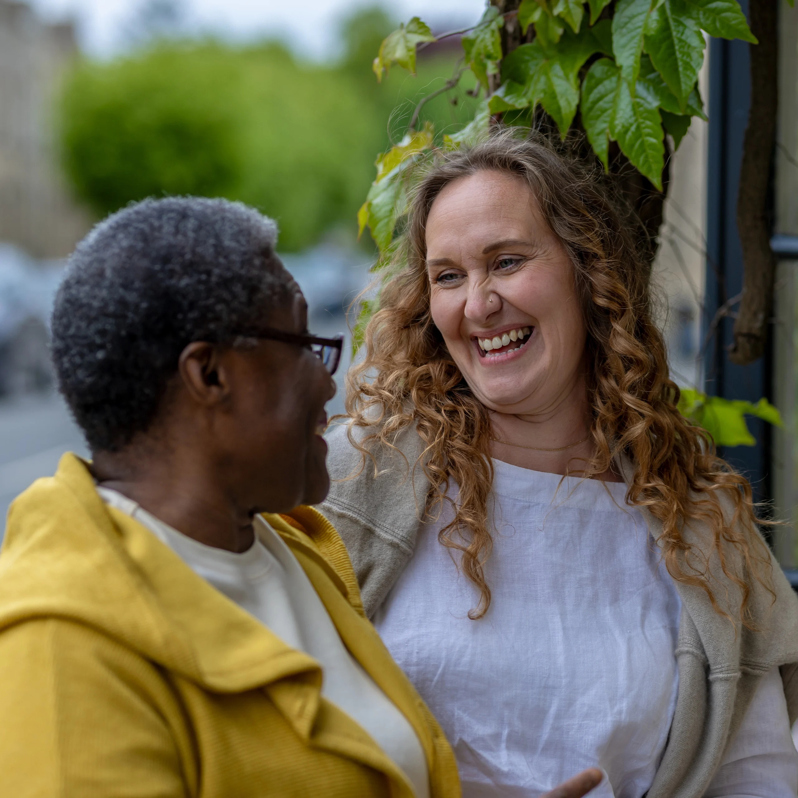 Two women smile and laugh together outdoors, standing close to each other near green leafy plants. One has curly hair and wears a white top; the other has short hair and wears glasses and a yellow jacket.