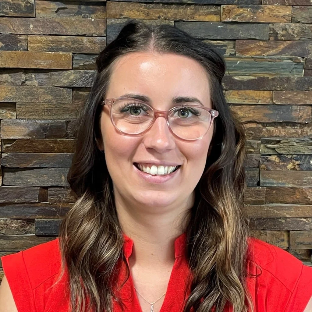 Meet our expert Lucy Childs: A woman with long brown hair, wearing glasses and a bright red top, smiles in front of a textured stone wall background.