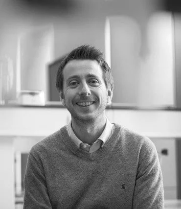 A man with short hair, wearing a collared shirt and a jumper, sits indoors and smiles at the camera. The image is in black and white.