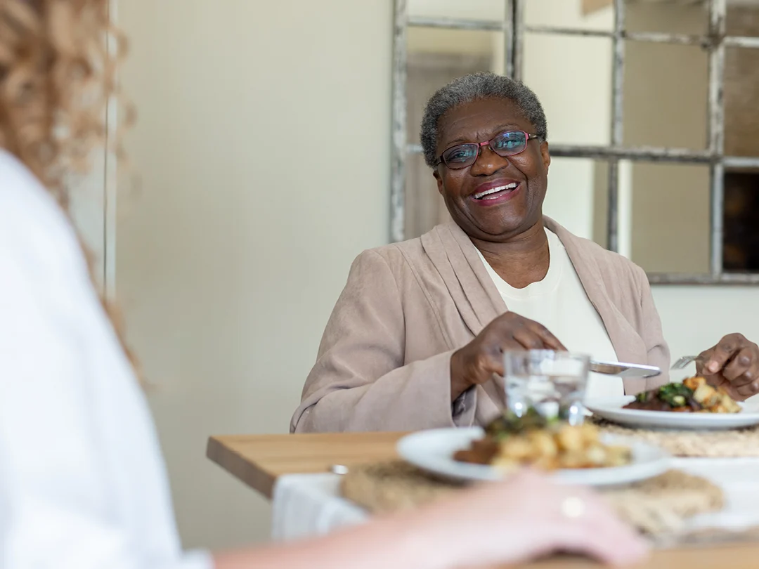 An older woman with short grey hair and glasses smiles whilst sitting at a dining table with a meal. Another person is seated opposite her, partially visible in the foreground.