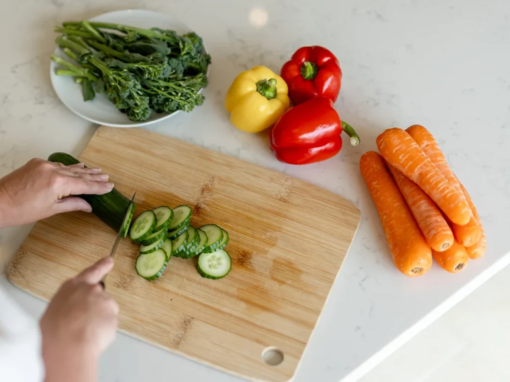 A person slices a cucumber on a wooden chopping board, surrounded by whole carrots, a red and a yellow pepper, and a plate of leafy broccoli on a white worktop.