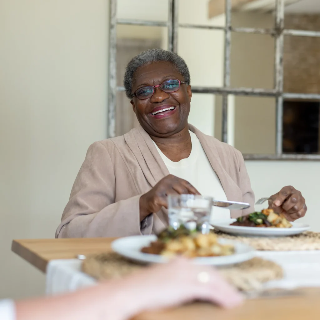 An older woman with short grey hair and glasses smiles while sitting at a dining table, holding a fork and knife, with plates of food and a glass of water in front of her.