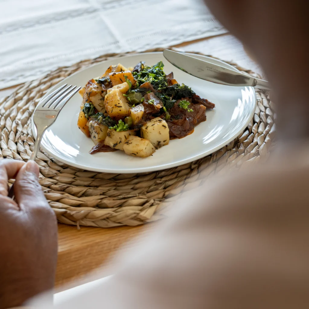 A person holding a fork and knife sits in front of a white plate with a meal of roasted potatoes, leafy greens, and vegetables on a woven placemat.