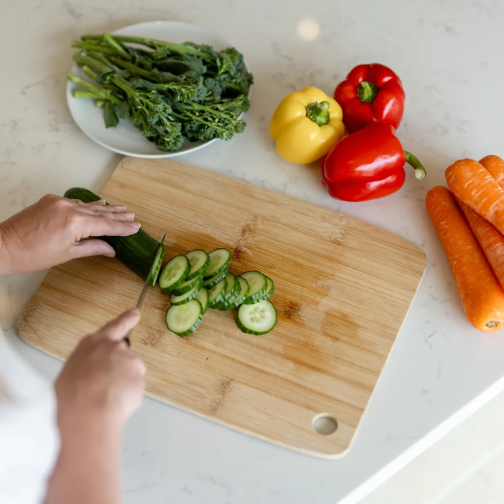 A person slices a cucumber on a wooden chopping board, surrounded by whole carrots, a yellow pepper, a red pepper, and leafy greens on a white worktop.