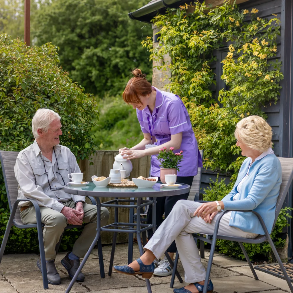 A carer in a purple uniform pours tea for an elderly man and woman sitting at a garden table, smiling and enjoying a sunny day. The table has tea cups, a teapot, and snacks. Greenery surrounds the patio area.