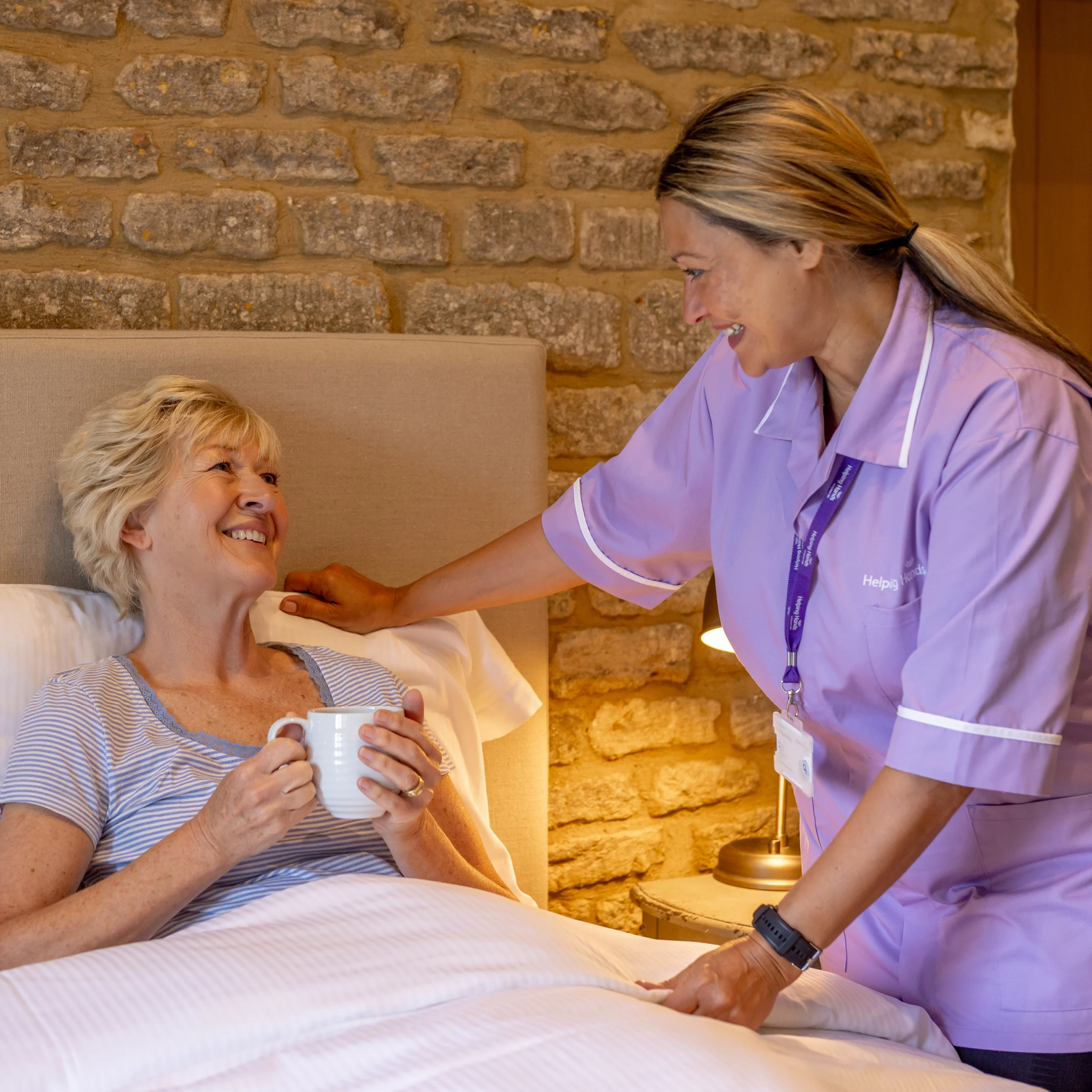 A carer in a purple uniform smiles and talks to an elderly woman sitting up in bed, holding a mug, in a cosy room with a stone wall background.