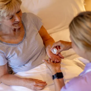 An older woman lies in bed, smiling, while a carer hands her medication tablets. The carer wears a smartwatch and both appear relaxed in a home or healthcare setting.