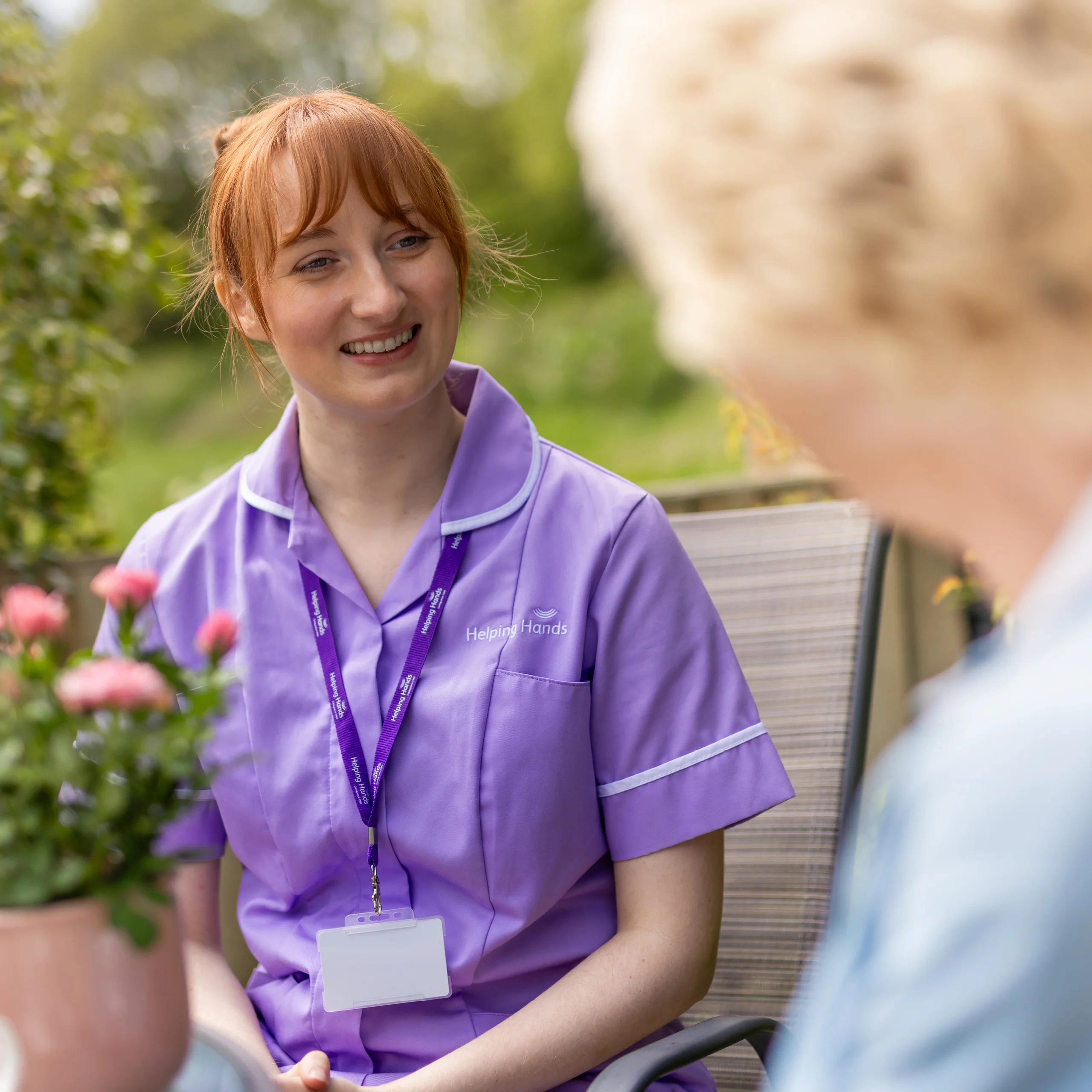 A smiling carer in a purple uniform sits outdoors, talking to an elderly person. The carer wears a lanyard and name badge, and pink flowers are visible in the foreground.
