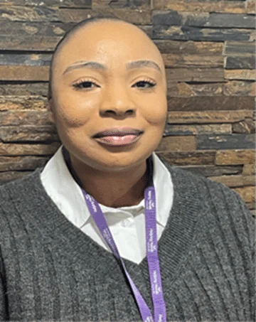 A woman with neatly styled hair smiles at the camera, wearing a white collared shirt under a grey jumper and a purple lanyard, standing in front of a textured stone wall.