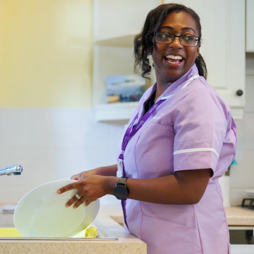A woman wearing glasses and a light purple uniform smiles while washing up at a kitchen sink. She is holding a white plate, and a yellow sponge is visible by the tap.
