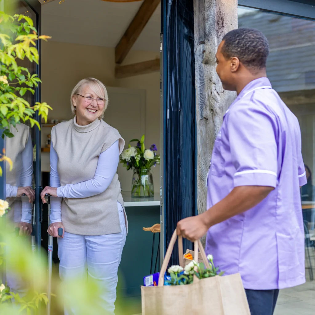 An elderly woman smiling at her open door as a carer in a purple uniform brings in a shopping bag filled with items. The scene is bright and welcoming, with flowers in the background.
