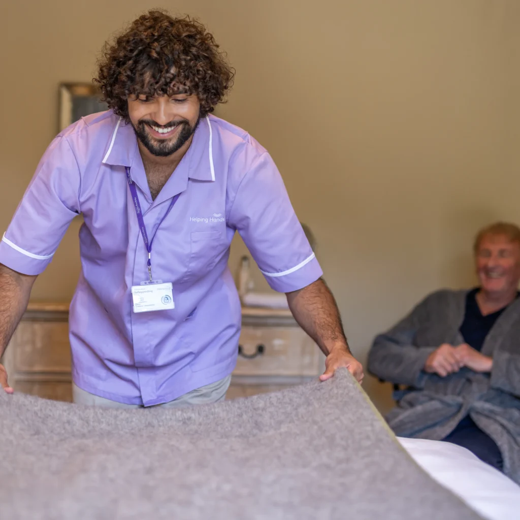 A smiling carer in a purple uniform makes a bed, while an elderly person in a dressing gown sits in the background, watching and smiling.