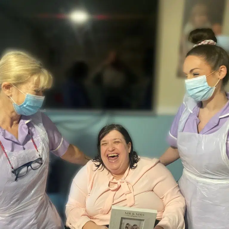 Three women stand together smiling; the woman in the centre is laughing joyfully while the two women on either side, both wearing face masks and aprons, look at her. The scene appears to be cheerful and supportive.