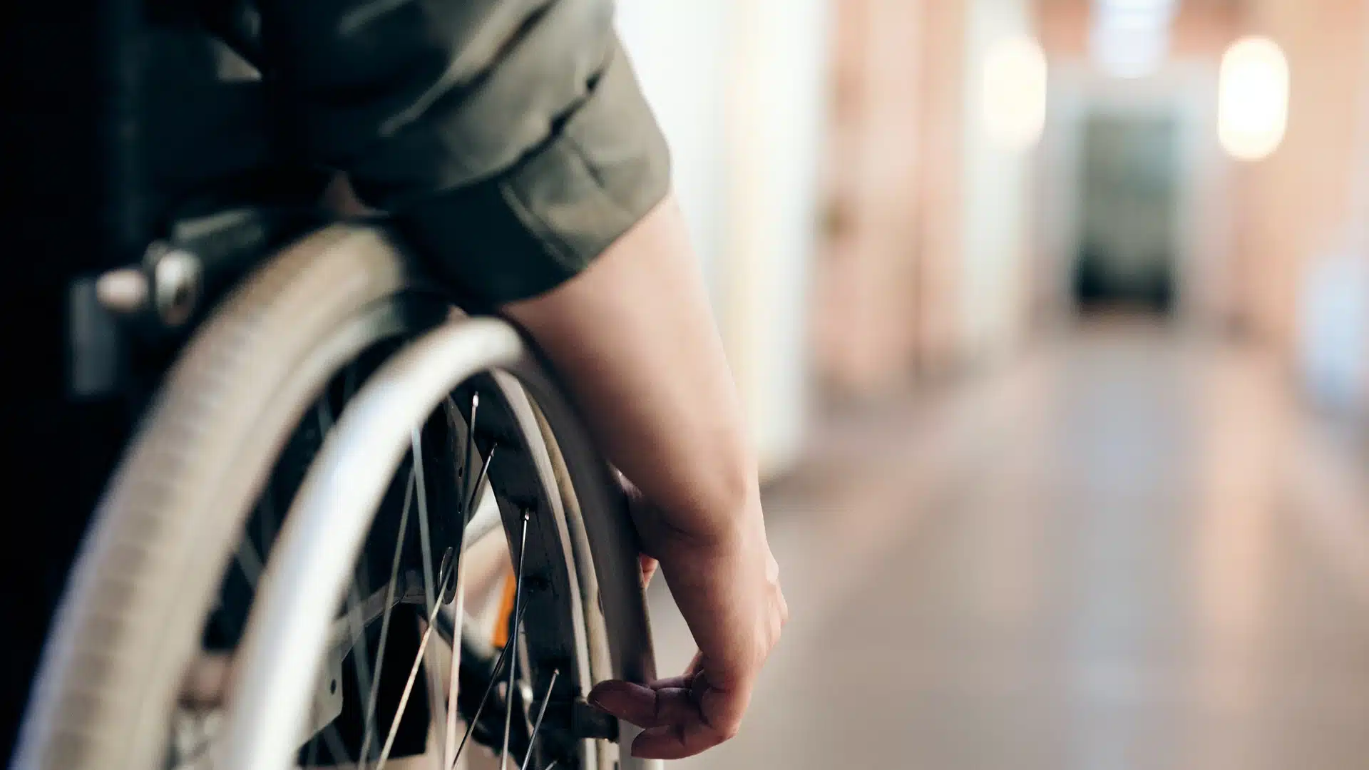 A close-up of a person’s hand on the wheel of a wheelchair, positioned in a corridor with a blurred background. The focus is on the arm and wheel, emphasising movement and accessibility.