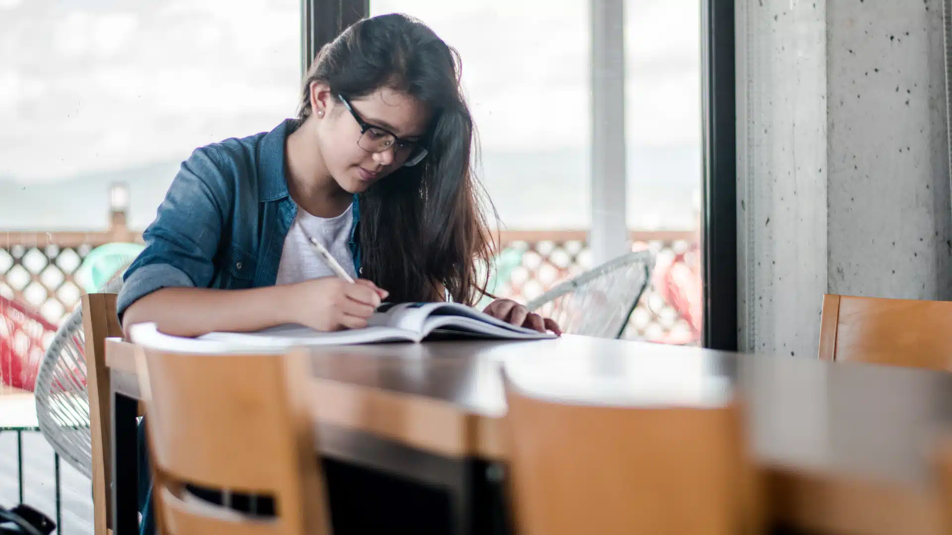 A young woman with glasses sits at a wooden table by a window, writing in a notebook. She appears focused and is surrounded by empty chairs in a bright, indoor space.