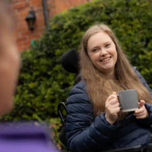 A woman with long hair, wearing a blue jacket, sits in a wheelchair and smiles whilst holding a mug. She is outdoors near green bushes, talking to another person whose face is blurred in the foreground.