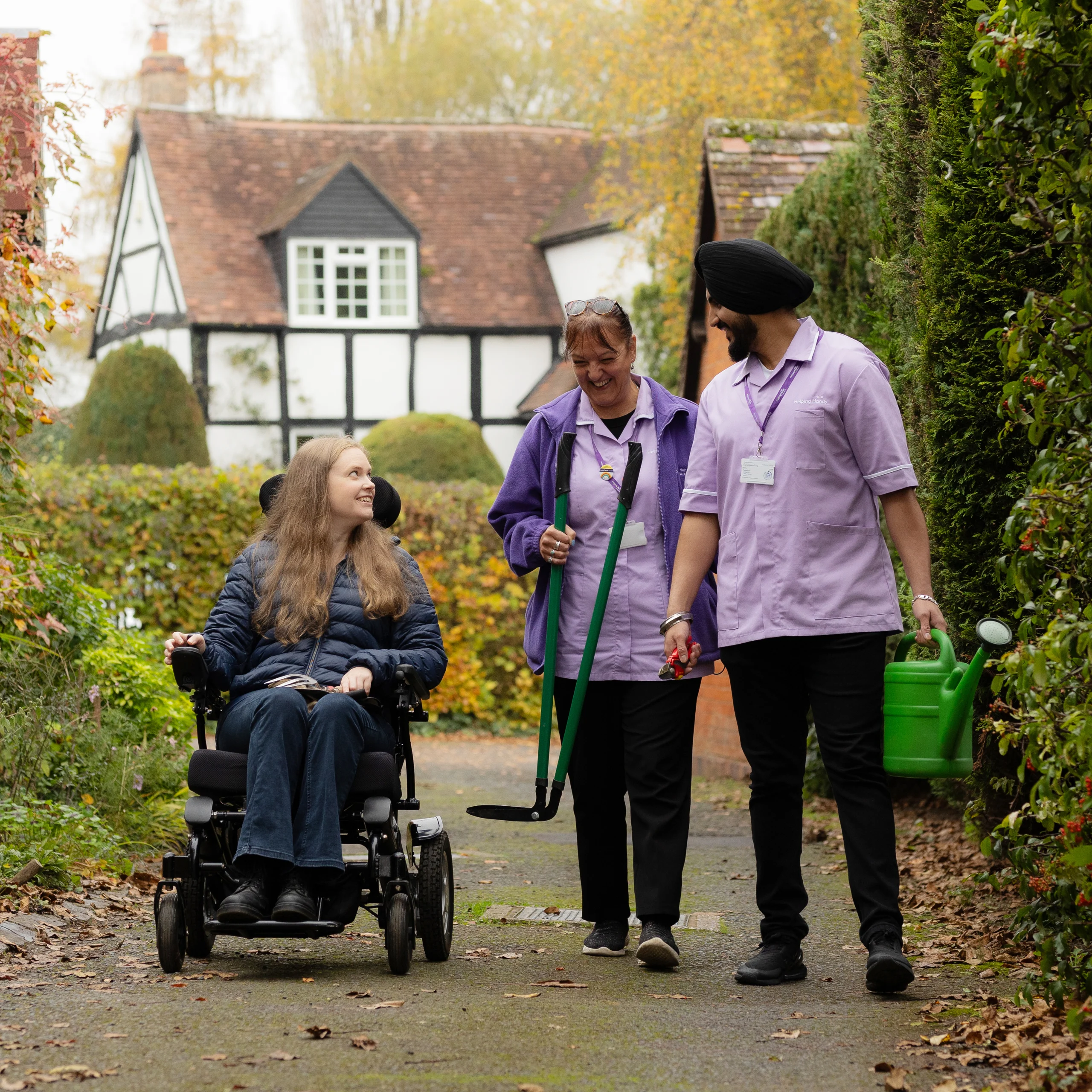 Two carers in purple uniforms walk outside with a young woman in a wheelchair on a path, holding gardening tools and a watering can, with a cottage and trees in the background.