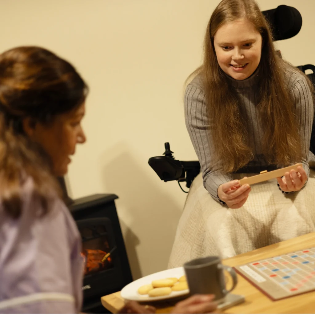 Two women play Scrabble at a table. One woman in a wheelchair holds letter tiles and smiles, while the other sits opposite her. There are biscuits, a mug, and a fireplace in the background.